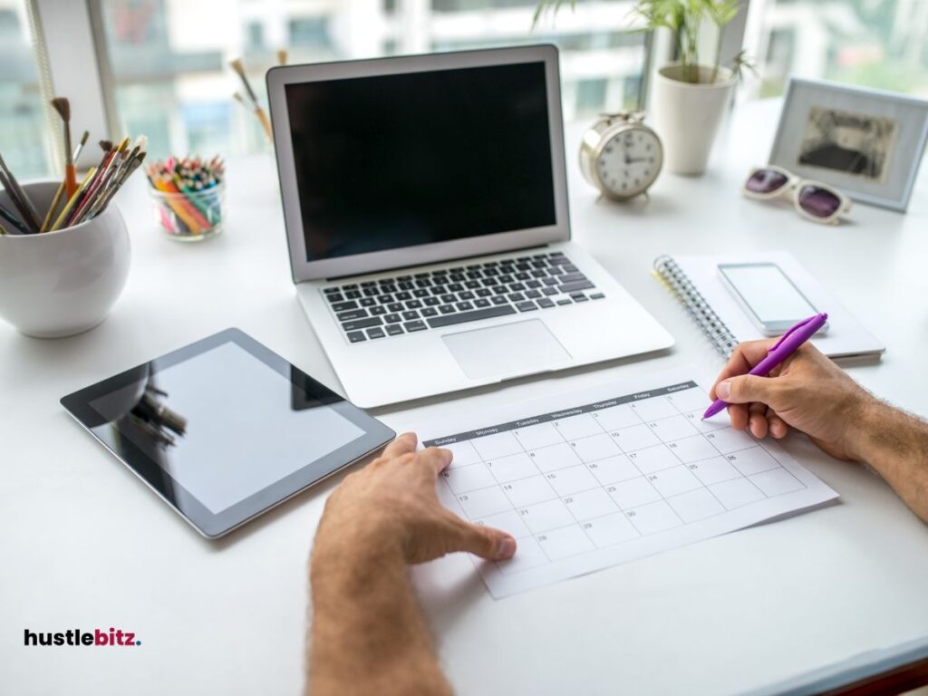 A hand of a man holding a calendar with a pen and a laptop wit tablet in the table