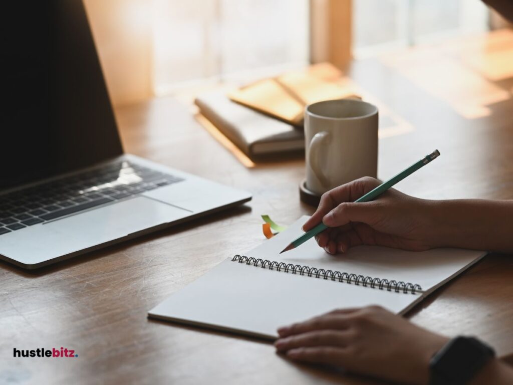 A hand doing writing on the notebook and a laptop in the table