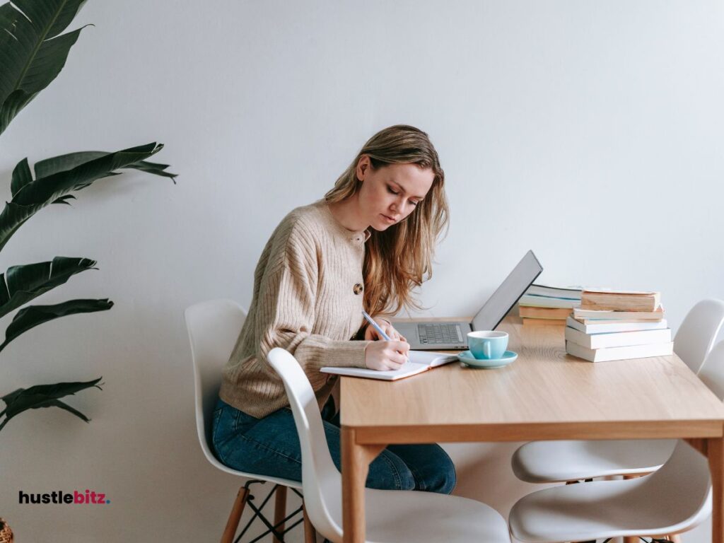 A picture of a woman writing on the notebook and a laptop in the table