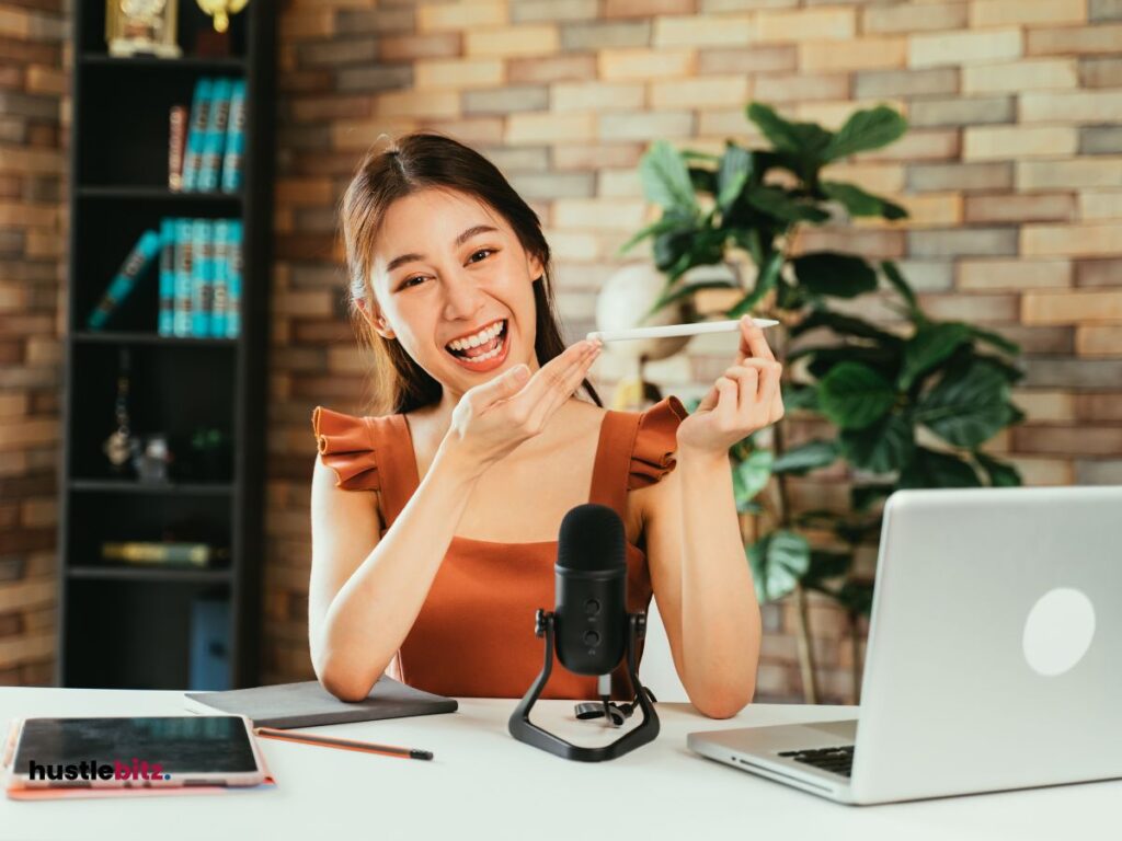 A woman smiles in front of the camera with laptop and mic in the table