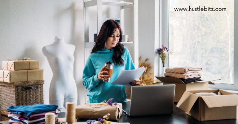 A woman in a blue hoodie holding coffee and papers, standing by a laptop in a workspace.