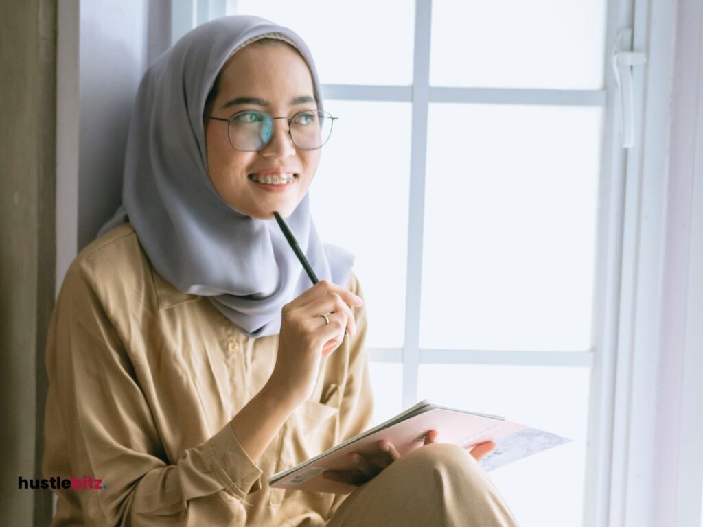 A woman sit beside the windows holding a pen and a notebook