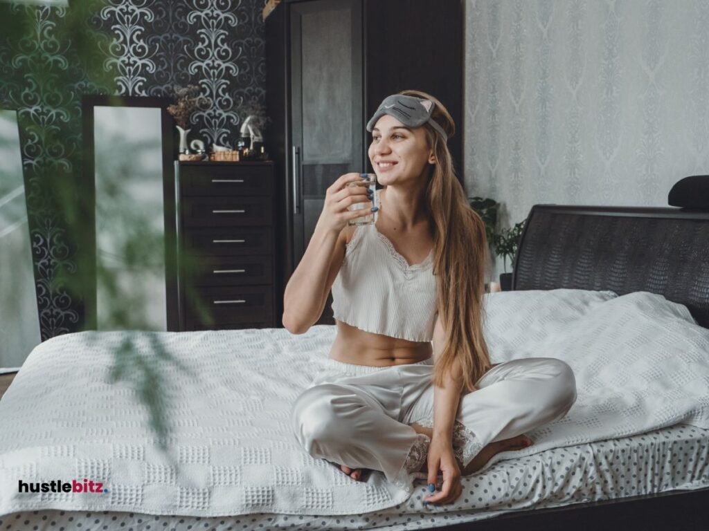 A woman holding a glass of water and smiles looking at the windows while sitting on the bed