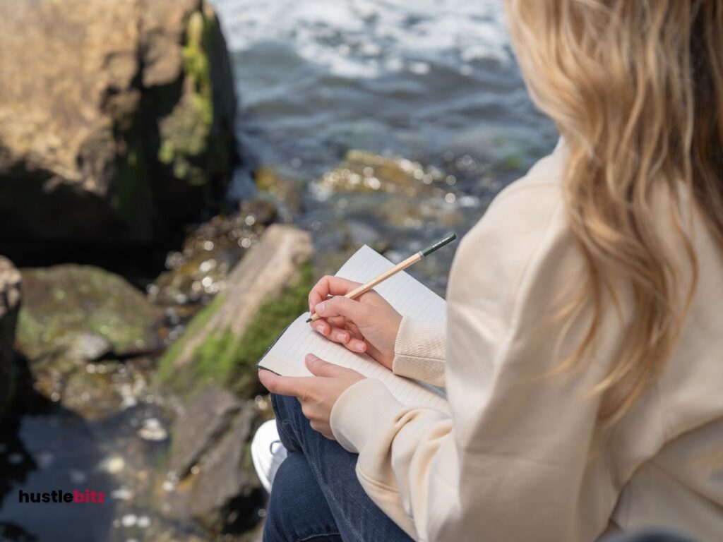 A woman holding a pen and a notebook sit beside the river