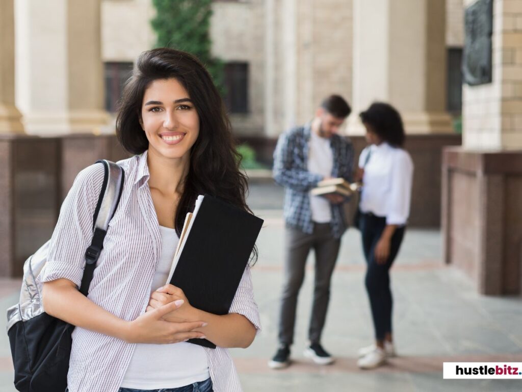 A smiling student holding books in front of a university, with two others talking behind her.