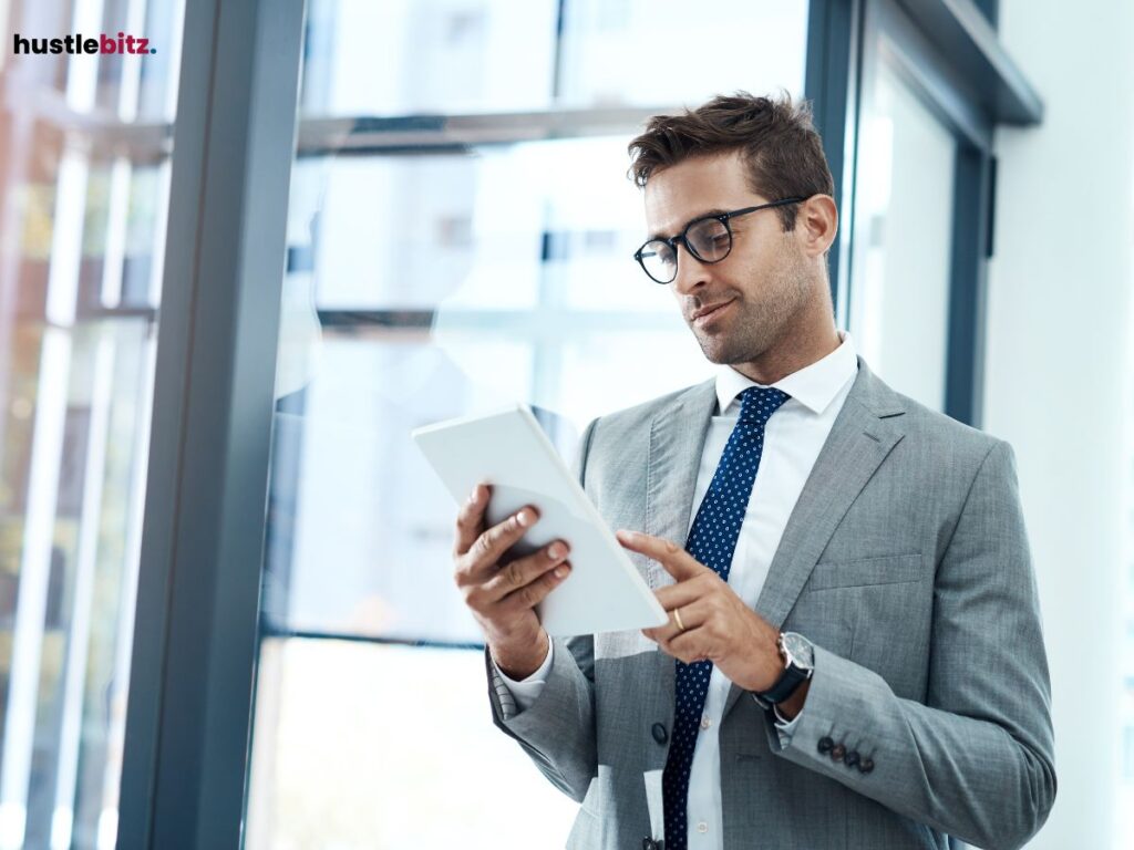 A man in a business suit and glasses, standing near a window, looking at a tablet.