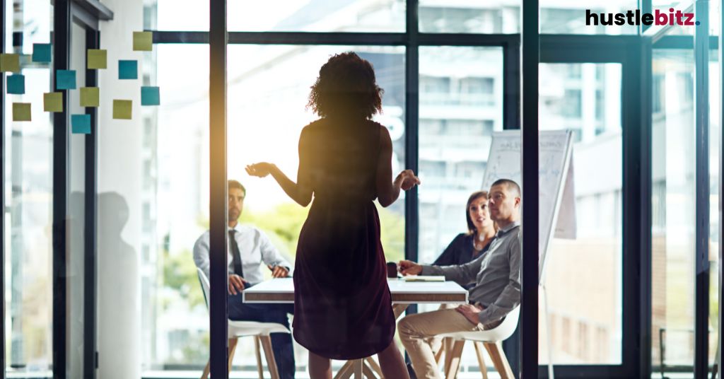 A woman leads a team meeting in a bright office with large windows.