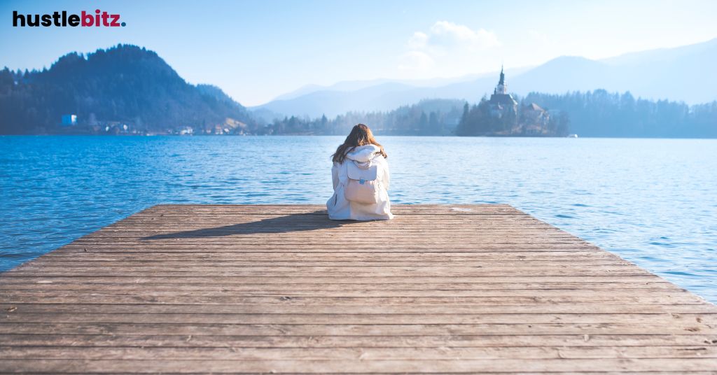 Person sitting on a dock, facing a peaceful lake with distant mountains.