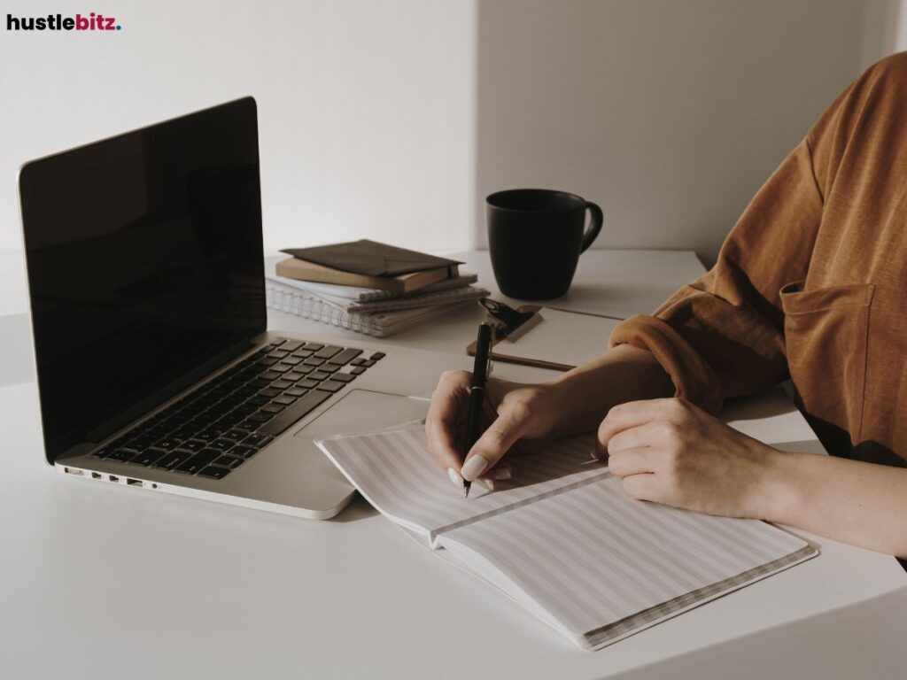 A person writing in a notebook at a minimalist desk with a laptop and books.