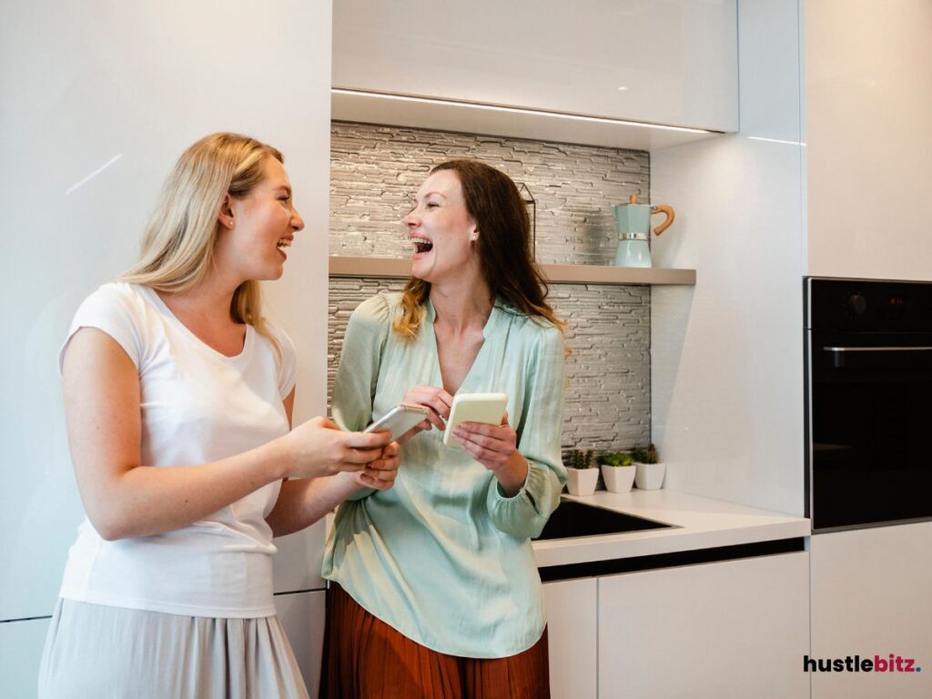 Two women laughing together while using their phones in a kitchen.