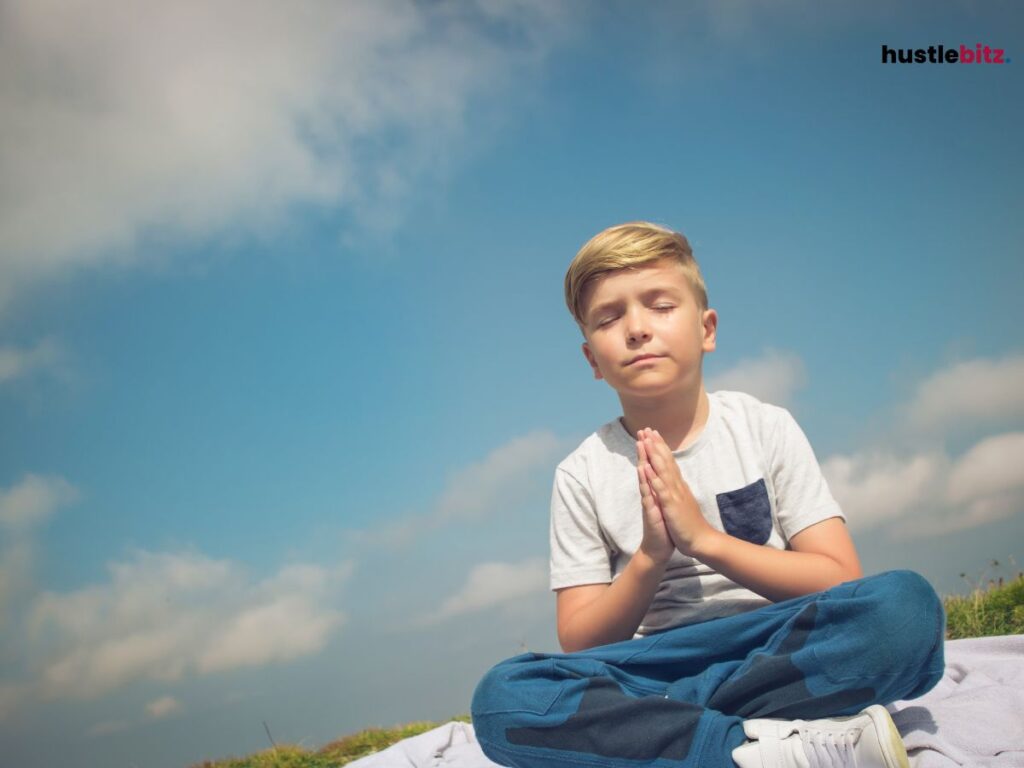 A young boy in a prayer pose sitting outside under a bright sky.