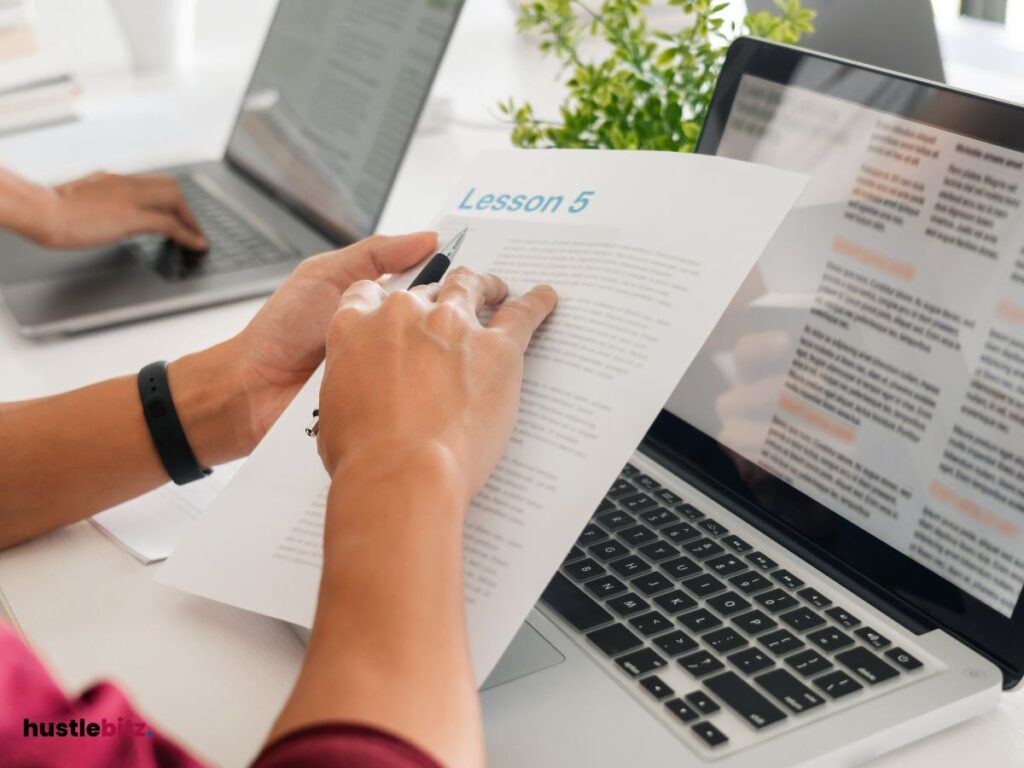 A hand holding a paper with pen and a table with laptop