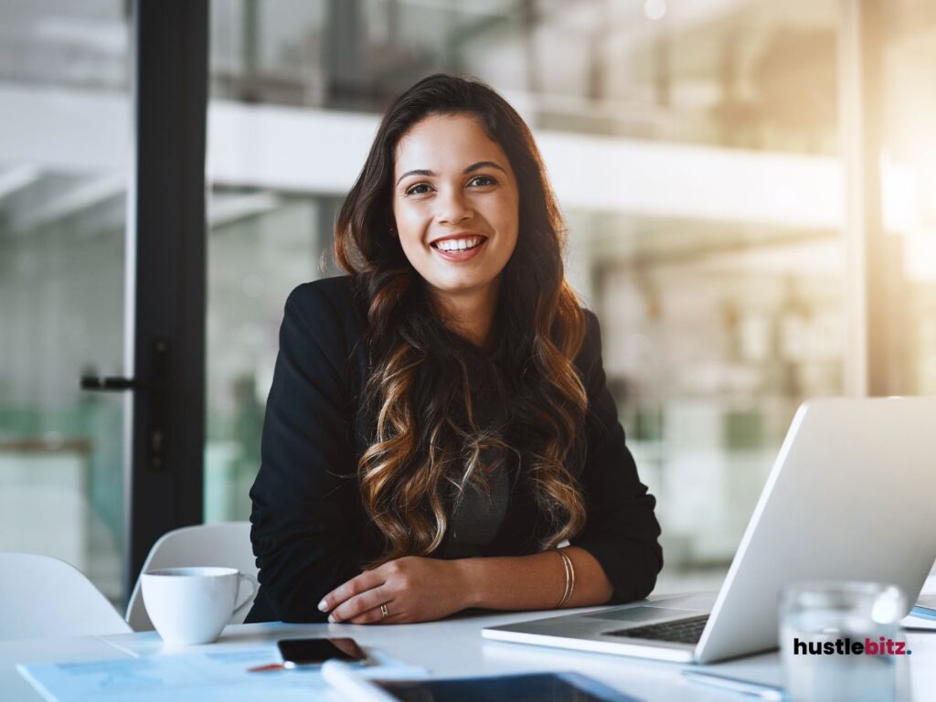 A woman smiles in front of the camera and a table with laptop