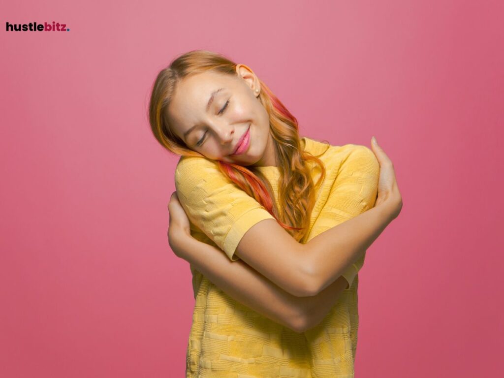 Smiling woman in a yellow shirt hugging herself against a pink background.