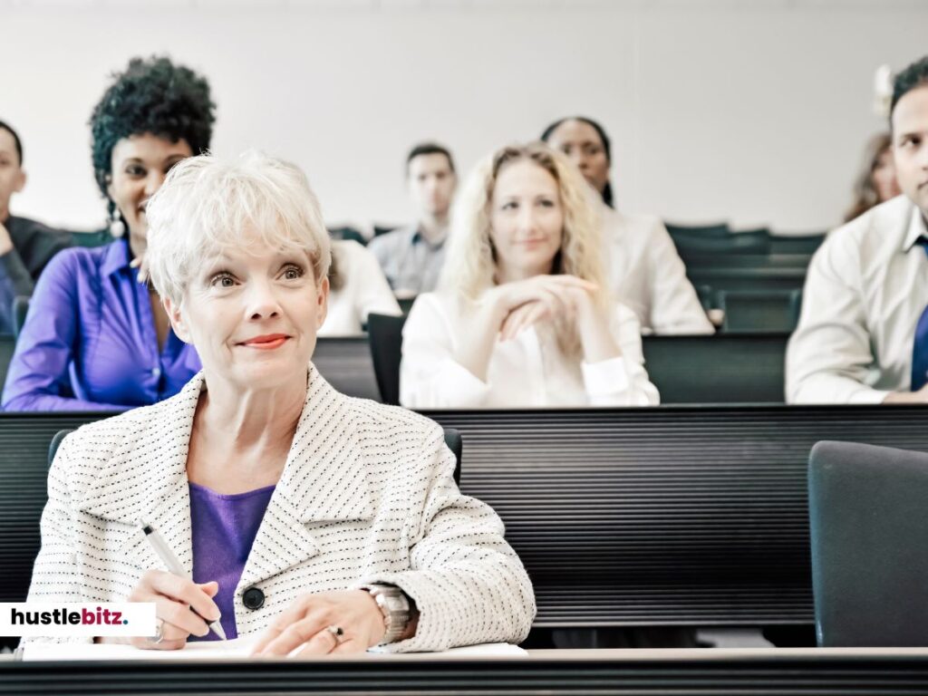 An older woman seated in a classroom setting, attentively taking notes while other are listening.