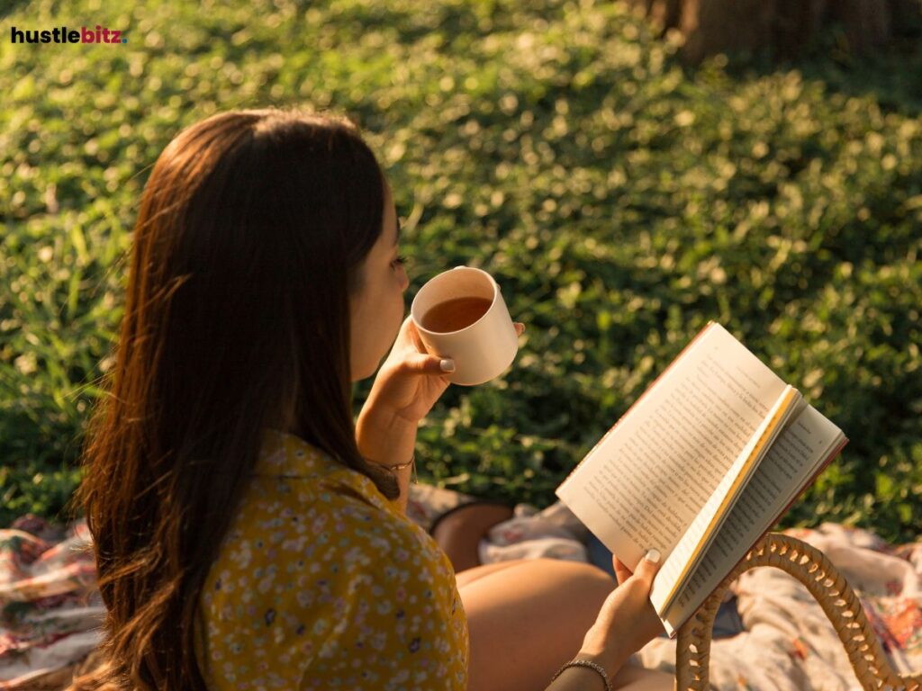 Woman sipping tea while reading a book outdoors on a grassy field.