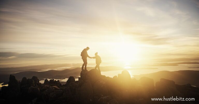 Two hikers on a mountain peak at sunrise, with one person helping the other up.