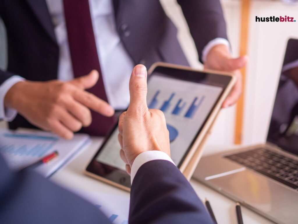 Businessmen giving a thumbs up and another person analyzing graphs on a tablet in a meeting.