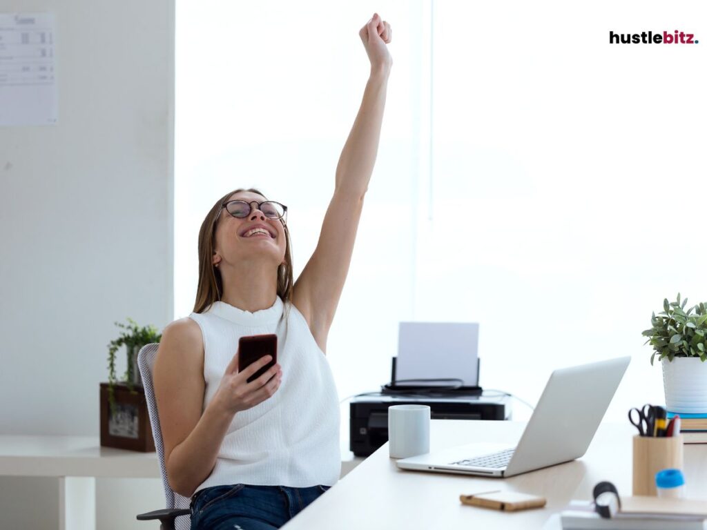  A woman at her desk, raising her arm in celebration while holding a phone.