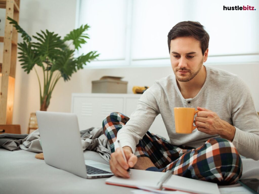 A man in pajamas, working with a laptop and notebook, holding a cup.