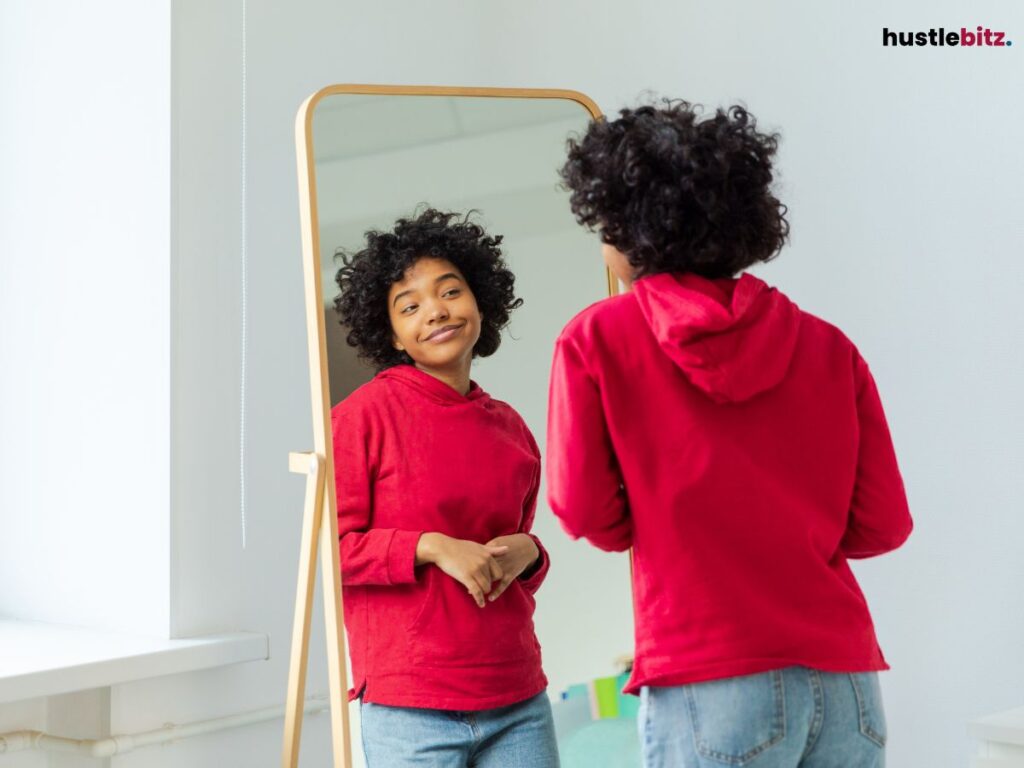 A woman smiling at her reflection in the mirror wearing a red hoodie.