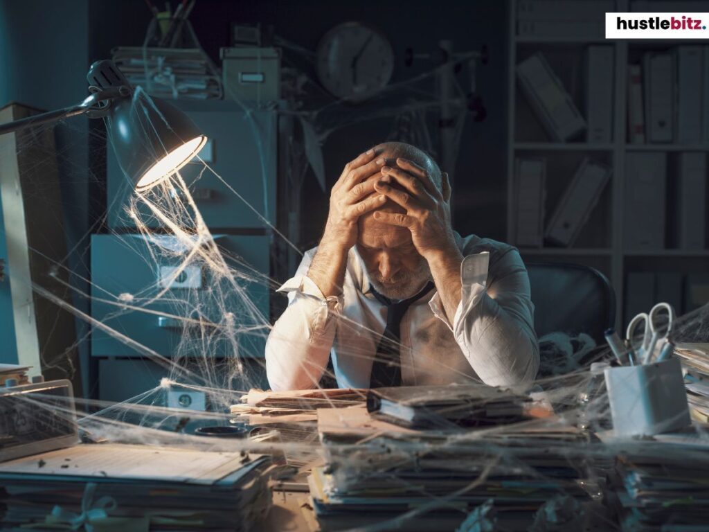 A man in a cluttered office with cobwebs, holding his head in frustration.