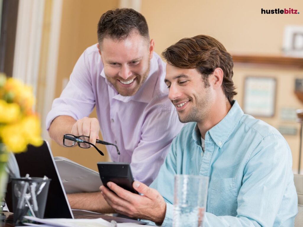 Two men smiling while collaborating over a phone and laptop.