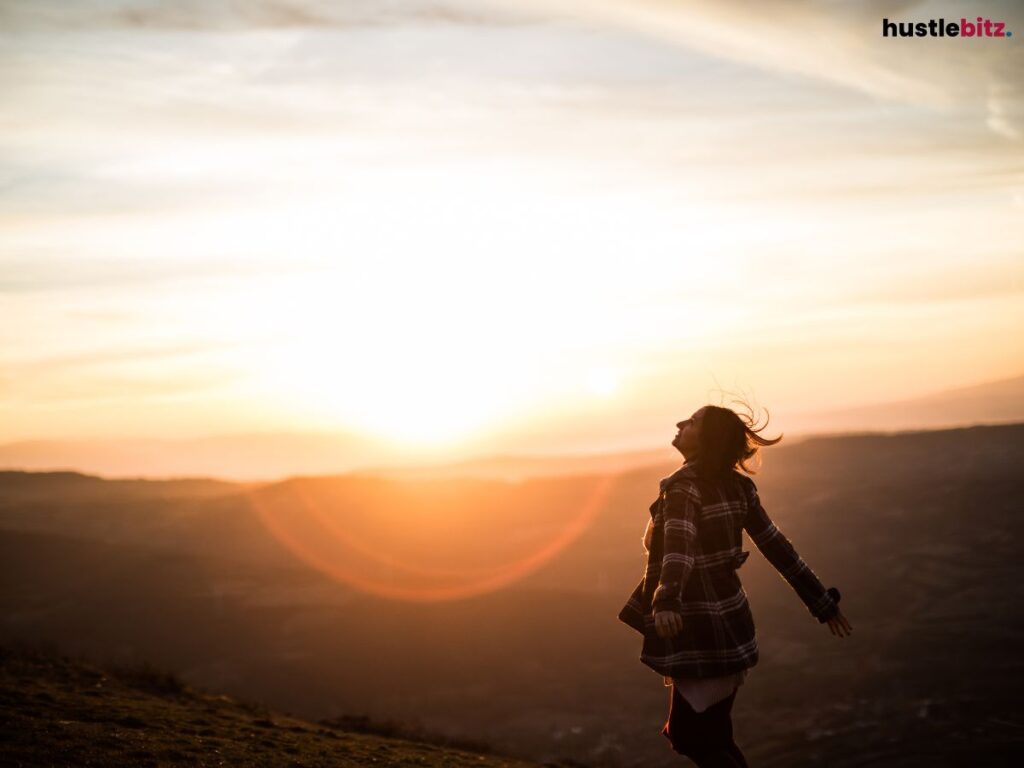 A woman with open arms enjoying a breathtaking sunset over a vast landscape.