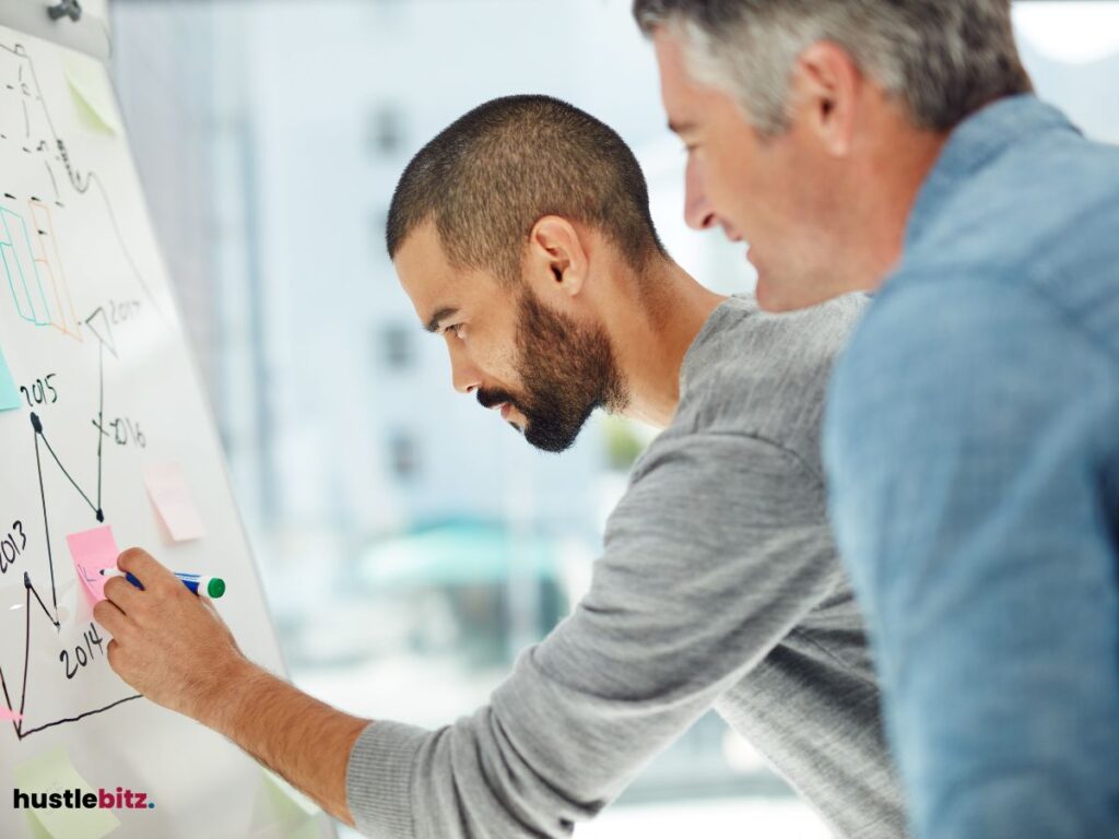 Two men collaborating at a whiteboard, with one drawing or writing while the other observes closely.