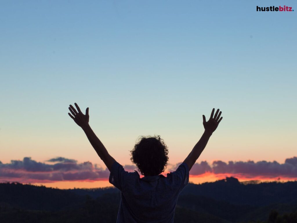 A silhouette of a person raising hands toward a sunset over mountains.