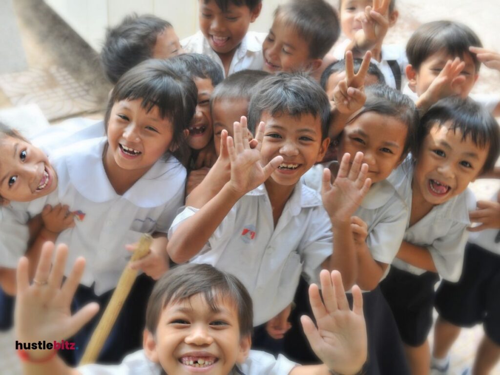 A group of smiling children in school uniforms enthusiastically wave at the camera.