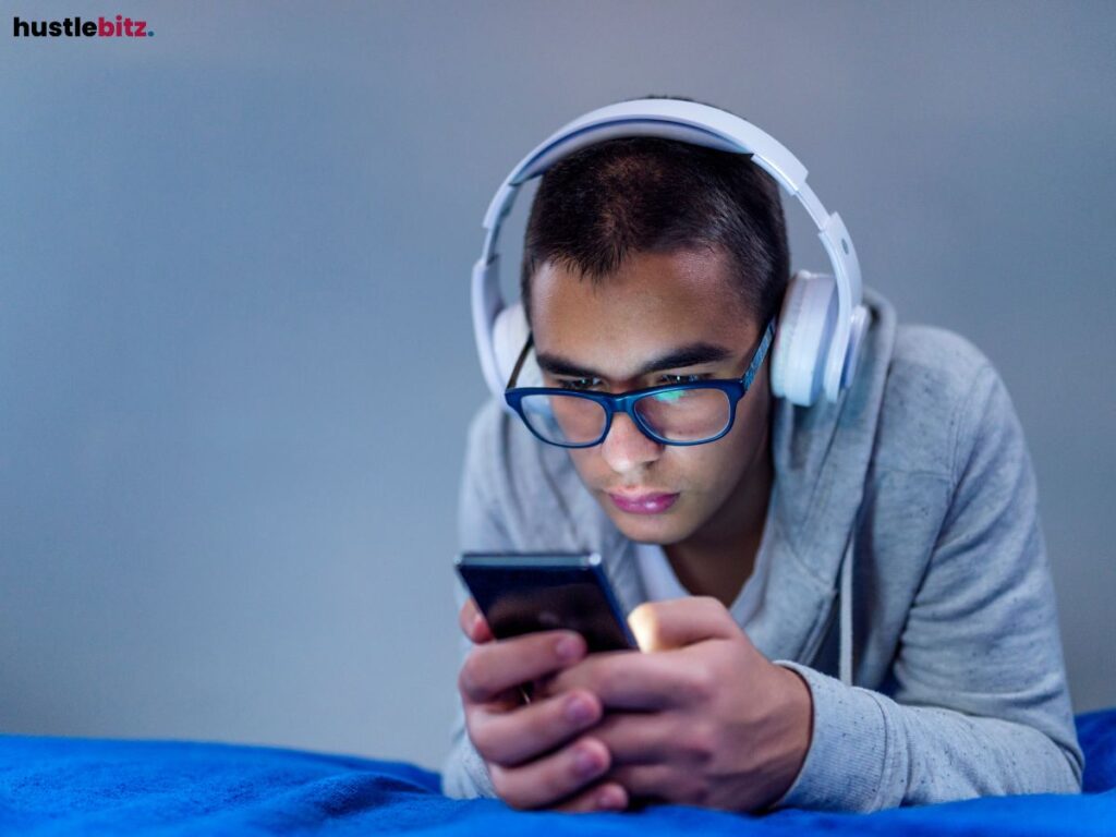 Man wearing headphones, focused on his phone, lying on a bed.