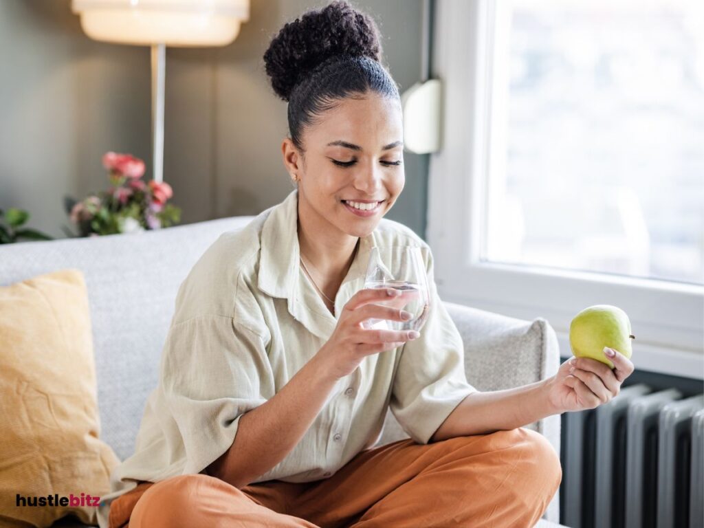 A woman relaxing at home, smiling while holding an apple and a glass of water.