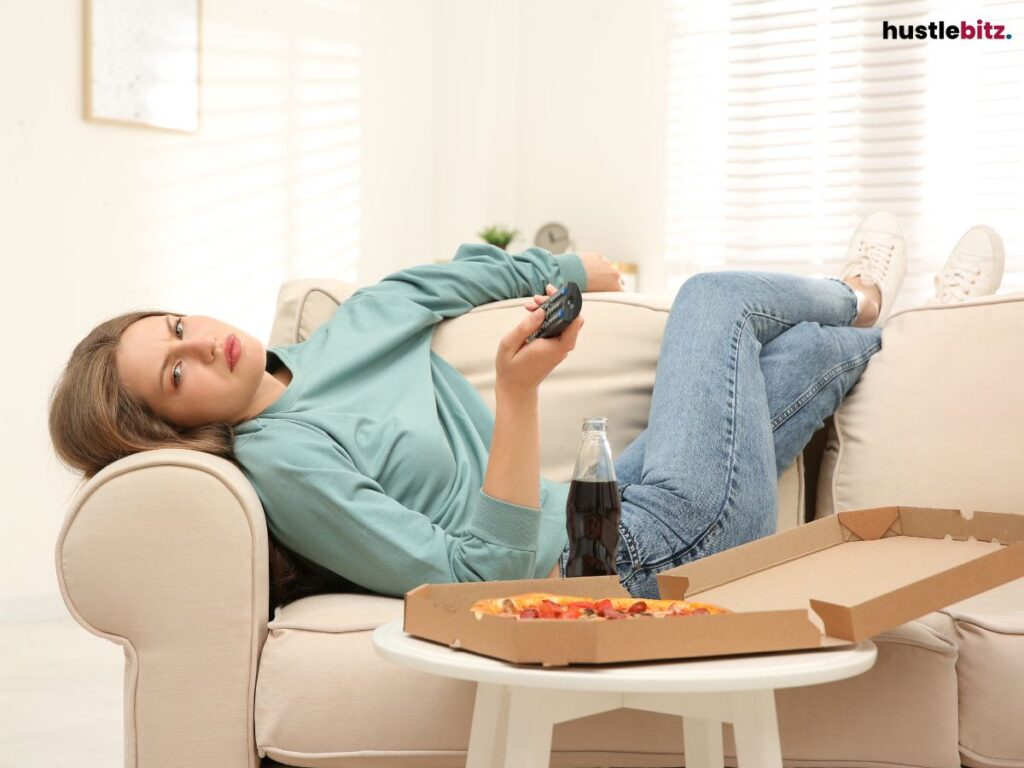 A woman lazily watching TV, surrounded by a soda and pizza box.