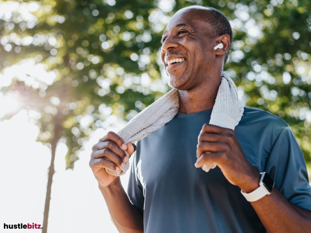Man smiling after exercising, holding a towel around his neck in a sunny park.