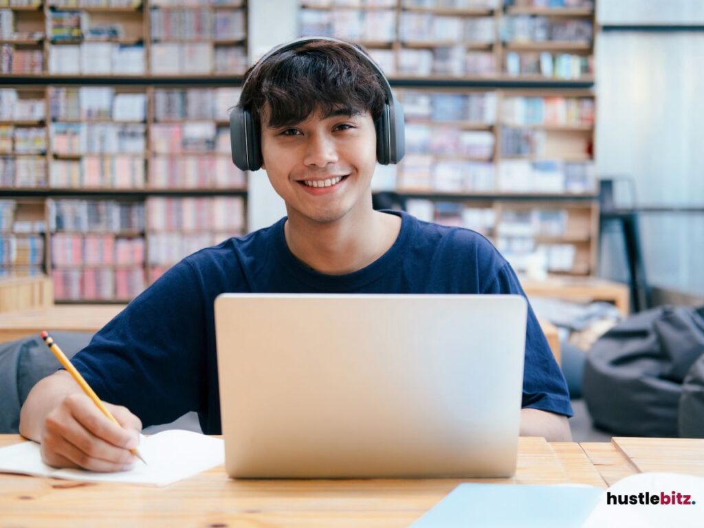 A young man wearing headphones smiles while studying with a laptop.