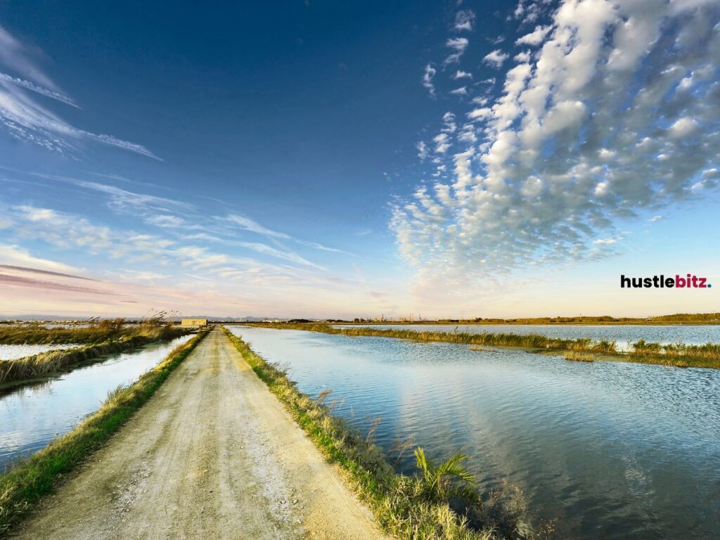 A long, straight dirt road flanked by bodies of water on both sides, extending toward.