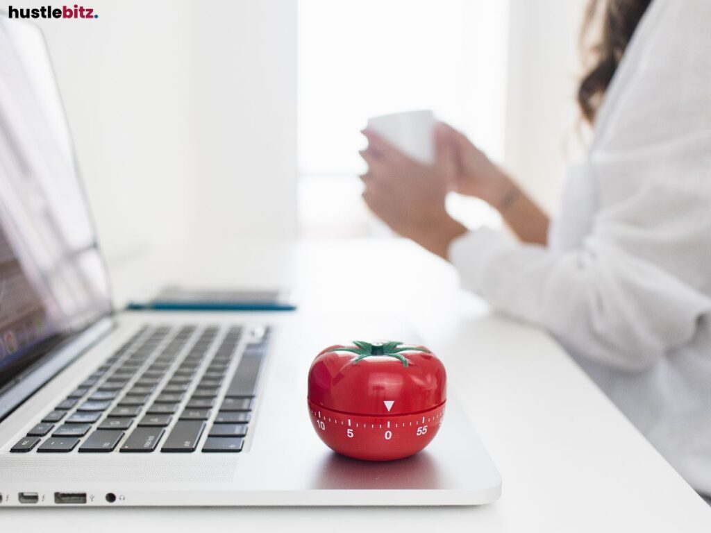 Laptop on a desk with a tomato-shaped timer and a person holding a mug.





