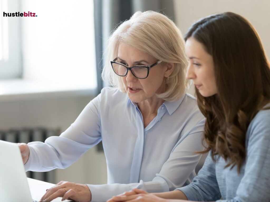 An older woman mentoring a younger woman, both focused on a laptop screen.