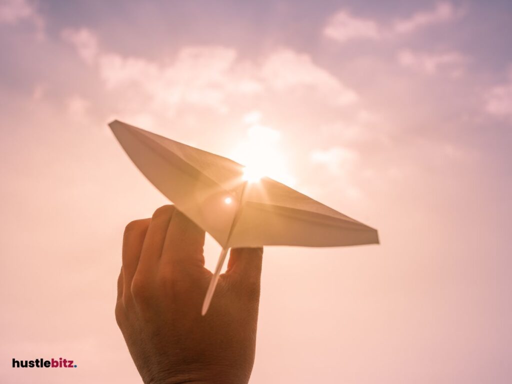 Hand launching a paper airplane into a bright, sunny sky.