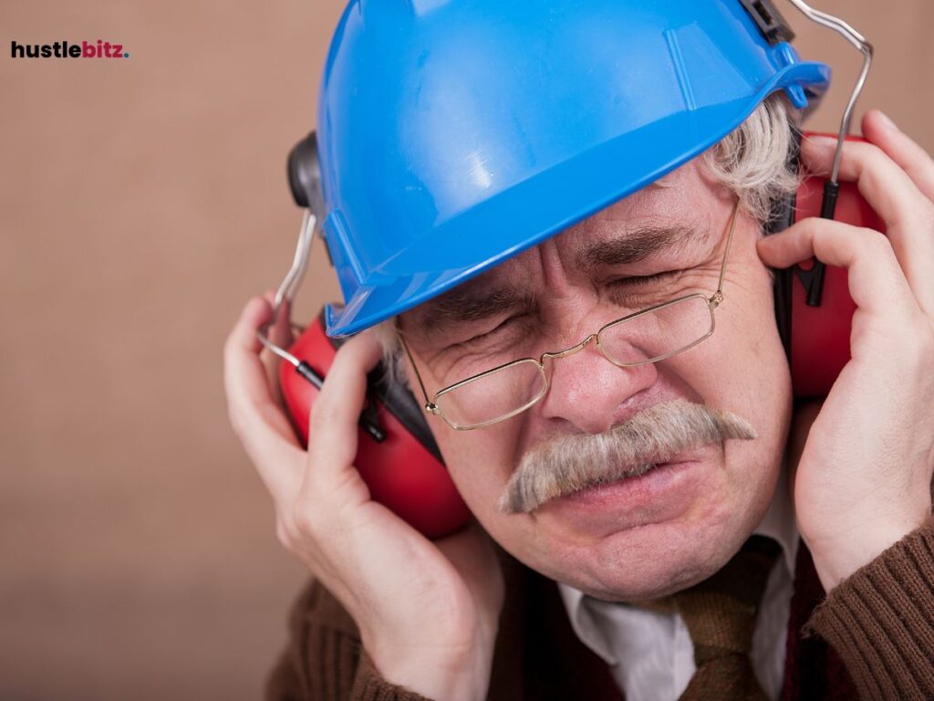 An older man in a hard hat covers his ears, grimacing from loud noise.