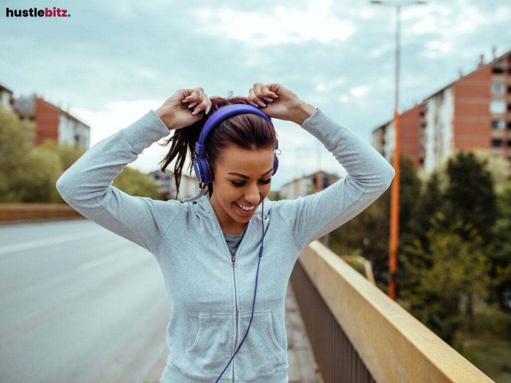 Woman outdoors, smiling, wearing headphones, and adjusting her ponytail.
