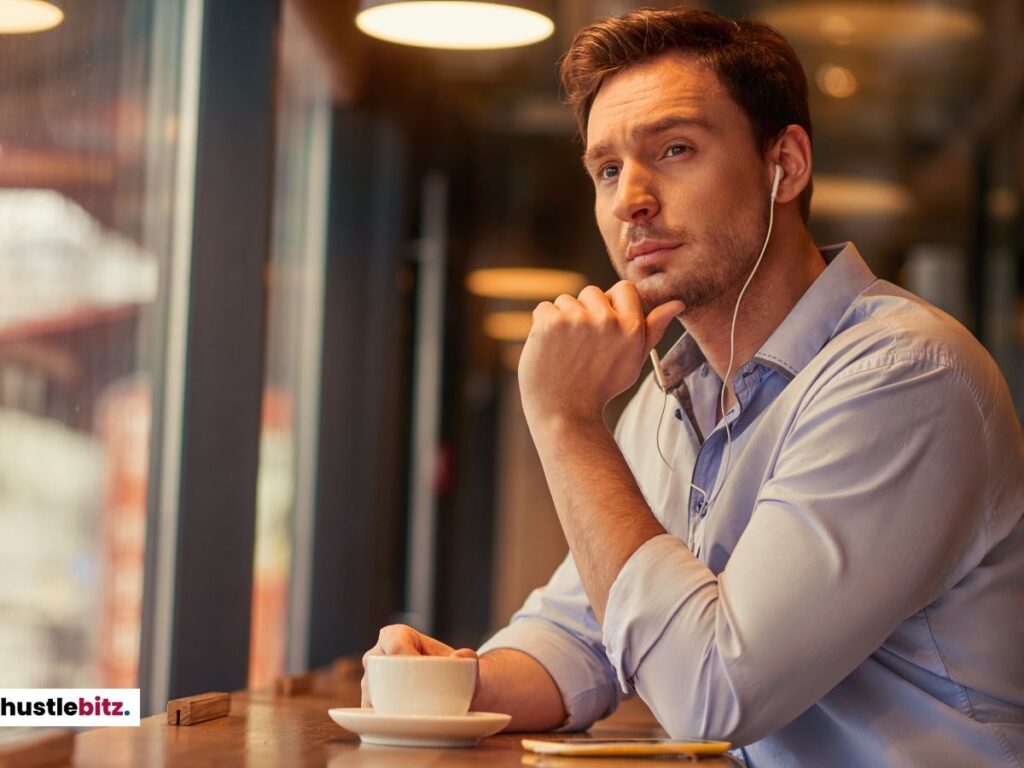 A man sitting by a window, wearing earphones, and gazing thoughtfully.