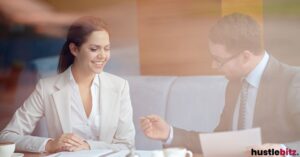 A smiling woman in a business setting discussing papers with a colleague.