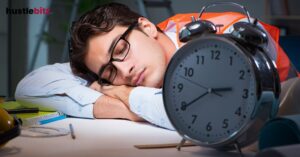 A man wearing glasses and an orange vest is napping at a desk, with a large clock nearby.