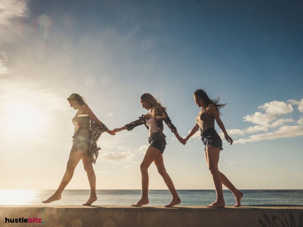 Three women walking barefoot, holding hands by the sea.