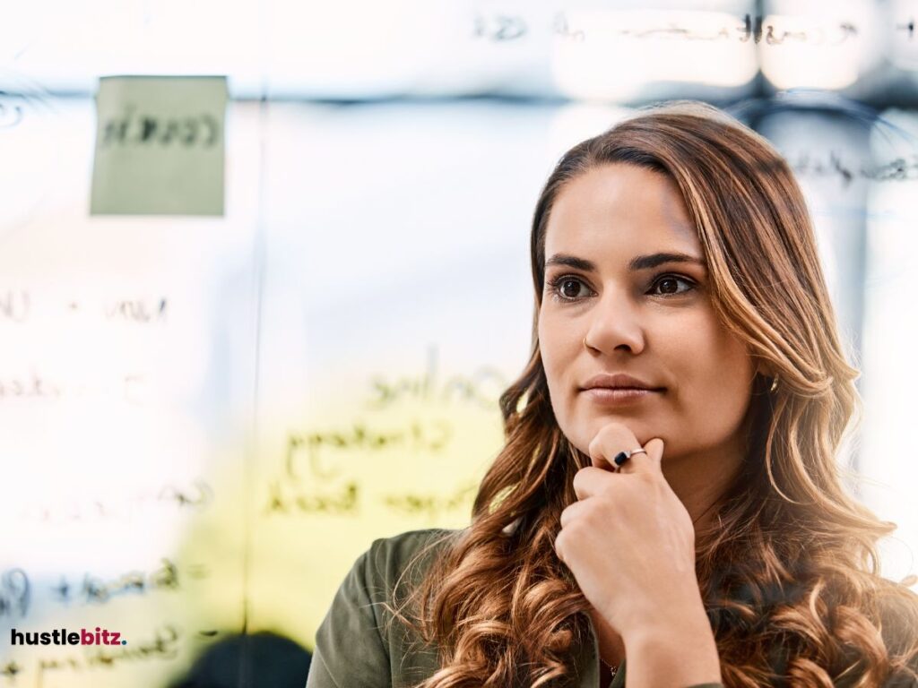 A thoughtful woman with her hand on her chin, looking at a glass board with notes.
