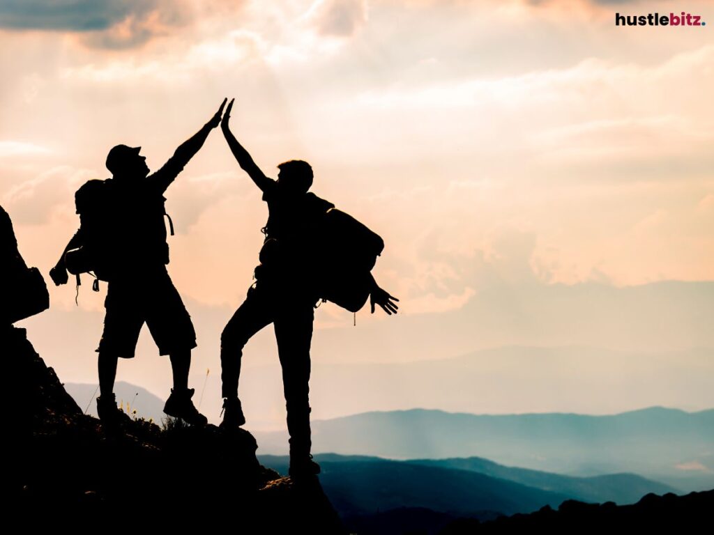 Silhouetted figures of two hikers high-fiving at the peak of a mountain.