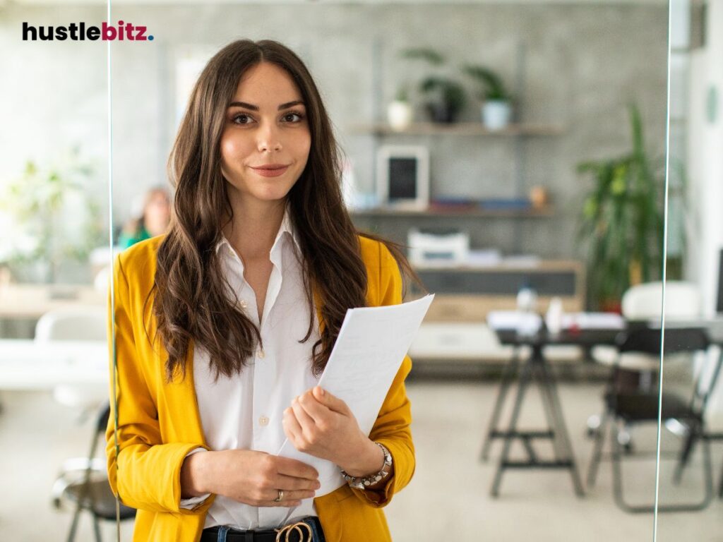 A confident woman in a yellow jacket holds papers, standing in an office.