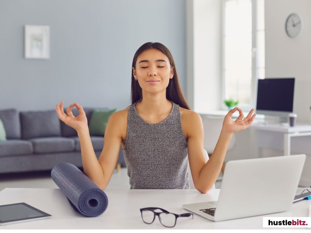 A woman meditating calmly at her desk with a yoga mat rolled up beside her.